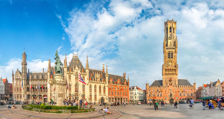 Brügger Marktplatz mit Belfried-Turm, historischen Gebäuden und Denkmal. Touristen versammeln sich auf dem Platz unter blauem Himmel.