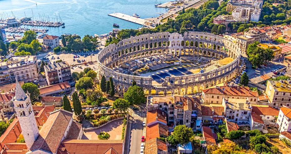 Luftaufnahme des antiken römischen Amphitheaters von Pula, umgeben von bunten Gebäuden mit roten Dächern. Der blaue Hafen mit Booten ist im Hintergrund sichtbar.