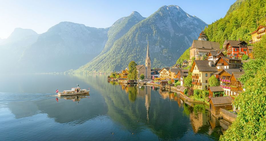 Hallstatt mit Kirchturm am ruhigen Seeufer, umgeben von bewaldeten Bergen. Ein Boot schwimmt auf dem spiegelnden Wasser.