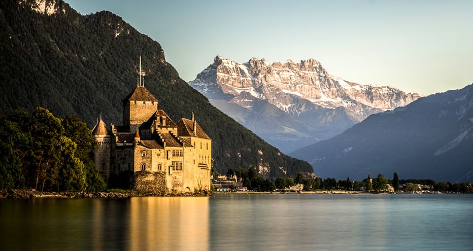 Schloss Chillon am Genfersee mit schneebedeckten Bergen im Hintergrund. Die mittelalterliche Festung steht im goldenen Licht am Wasser.