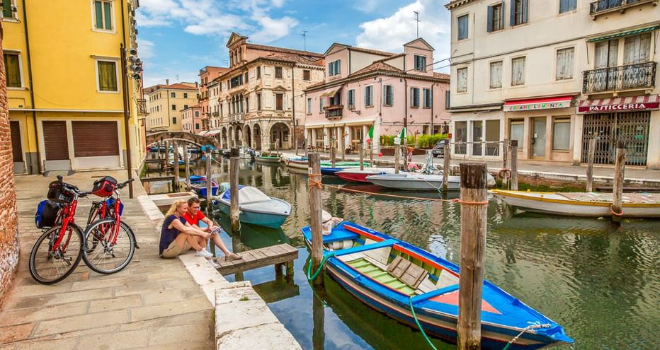 Radfahrer mit roten Tourenrädern rasten an einem Kanal in einer italienischen Stadt. Bunte Boote liegen an Holzpfählen vertäut, historische Gebäude säumen den Kanal.